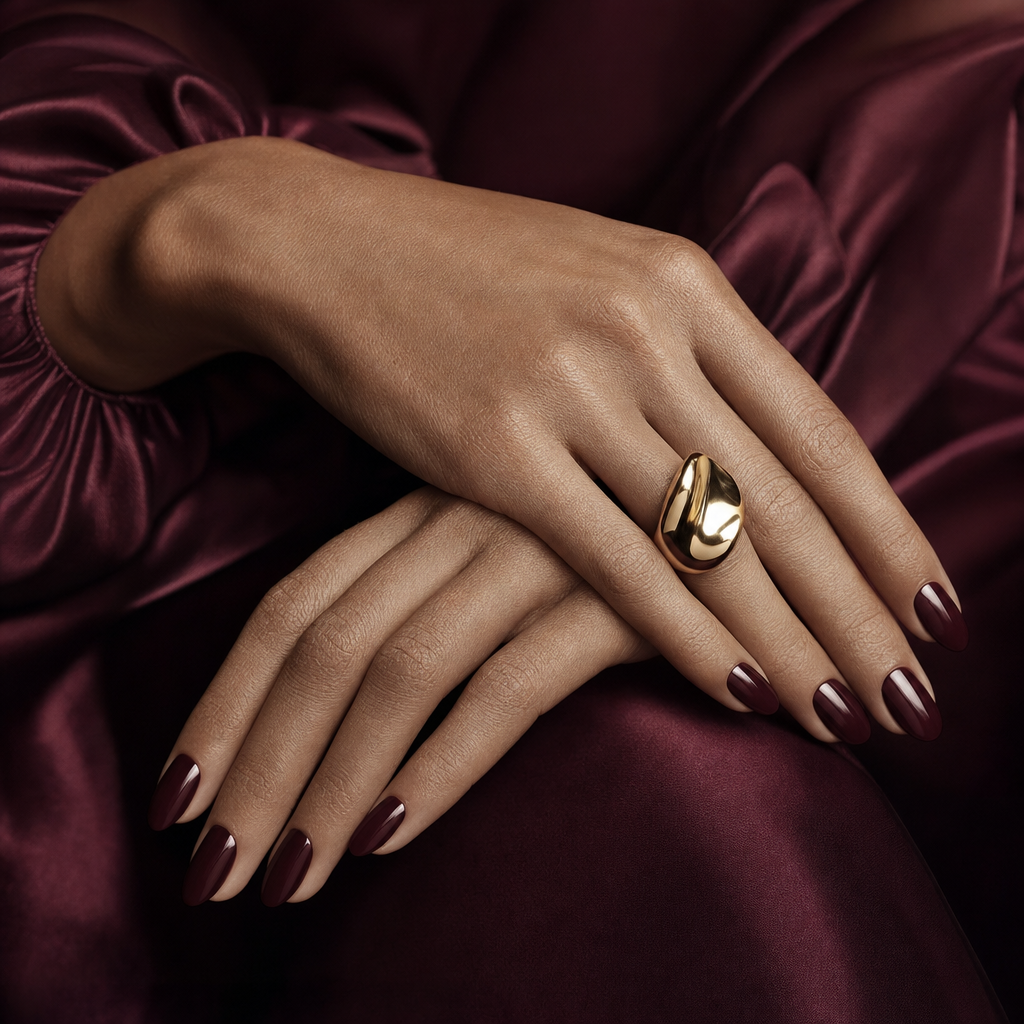 Editorial macro of elegant hands with oxblood manicure and a sculptural gold ring on draped silk